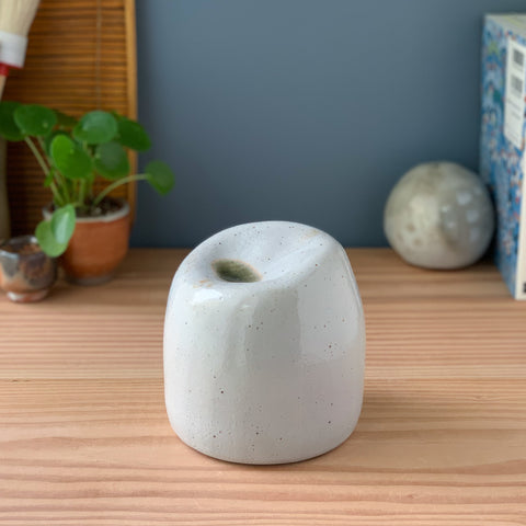 A white glazed closed-form ceramic sculpture. It has a sloped indentation on the top. Inside the indentation is a pool of glassy green glaze. The sculpture sits on a wood table with a plant and books in the background.