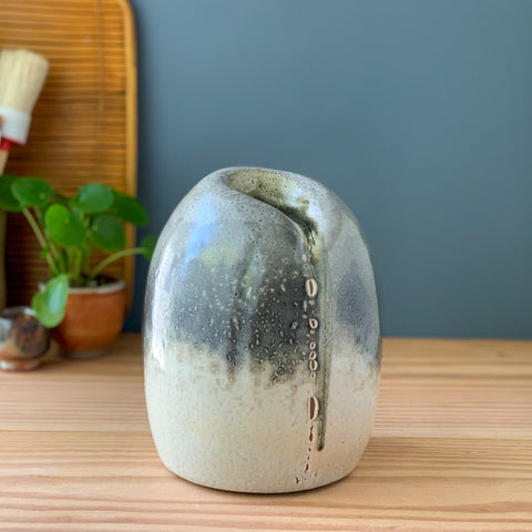 Abstract ceramic closed-form sculpture with grey and white crawling glaze on a wood table.